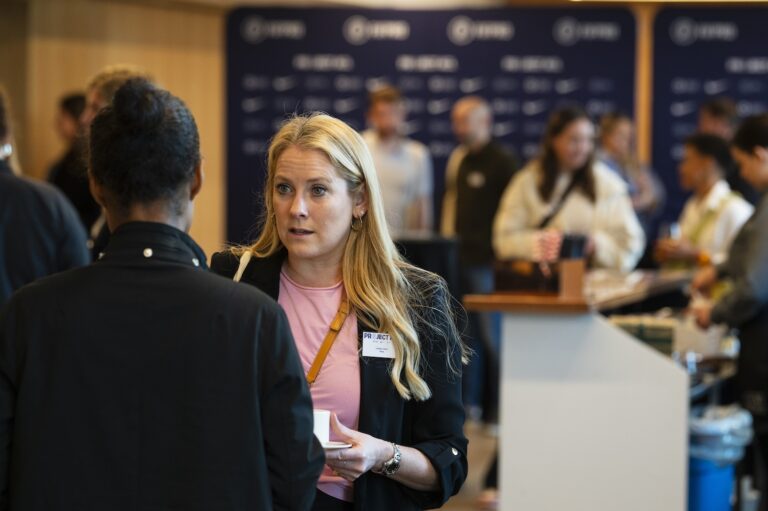 Two women having a conversation in a busy breakout room. Behind them are people mingling.